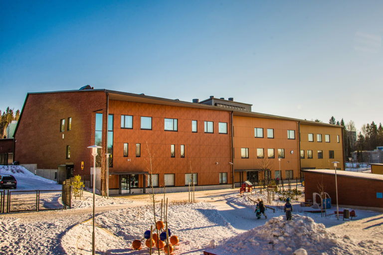 Liipolan monitoimitalo kuvaa, kuvassa näkyy Trespa asennus - kolme eri väristä seinää vieri vieressä, tumman ruskeasta kellertävvään puukuvioon. Etualalla leikkipuisto. Julkisivuremontti.